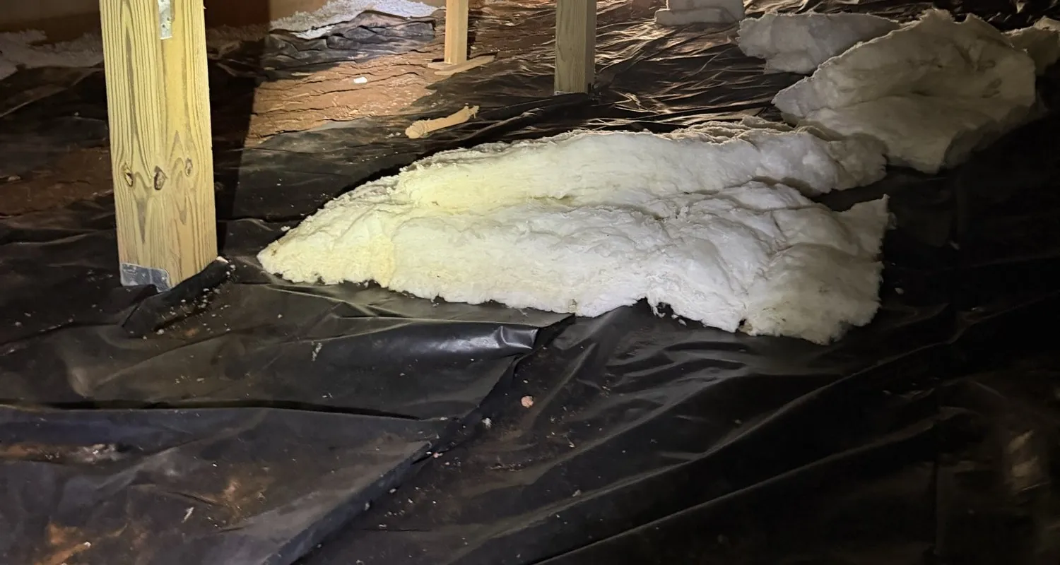 A crawl space before treatment: insulation fallen onto torn black plastic between the wood posts